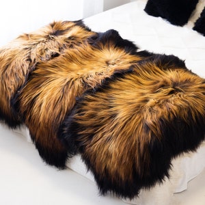 May include: Three sheepskin rugs in shades of brown and black, arranged on a white bed. The rugs have long, fluffy fur.