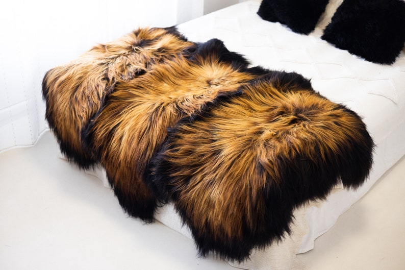 May include: Three sheepskin rugs in shades of brown and black, arranged on a white bed. The rugs have long, fluffy fur.