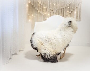 May include: A white sheepskin rug draped over a white rocking chair in a white room with a macrame wall hanging and string lights.
