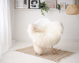 May include: A white sheepskin rug draped over a white chair with wooden legs. The chair is sitting on a woven rug with a light brown and beige pattern.