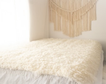May include: A white fluffy rug or blanket on a white bed with a white wall and a macrame wall hanging in the background.