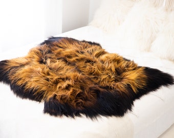 Op de afbeelding: Een bruin en zwart schapenvacht met lange, zachte vacht. Het kleed ligt op een wit beddengoed.