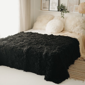 May include: A black fluffy throw blanket on a bed with white pillows. The blanket is made of a soft, plush material and has a shaggy texture.