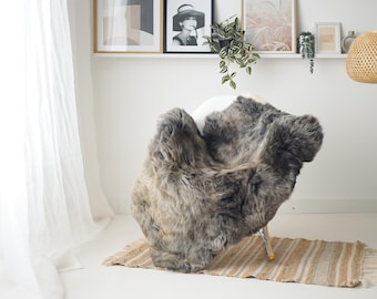 Può includere: Un tappeto in pelle di pecora grigio e marrone drappeggiato su una sedia bianca. Il tappeto è posto su un tappeto di iuta a righe. La stanza ha un'estetica minimalista con opere d'arte incorniciate e una lampada a sospensione intrecciata.