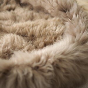 May include: Close-up of a soft, fluffy, light brown faux fur fabric. The fur has a dense, textured appearance with long, flowing fibres. The edges are slightly lighter in colour, creating a subtle contrast. The fabric appears to be a rug or throw.