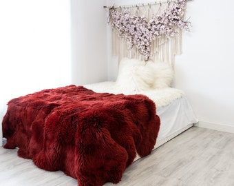 May include: A red faux fur throw blanket on a white bed with a white faux fur pillow. A macrame wall hanging with pink flowers is above the bed.