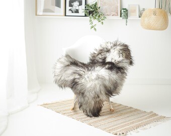 May include: A white chair with a sheepskin throw draped over it. The sheepskin is white and gray with long, fluffy fur. The chair is sitting on a light brown rug with a fringe.