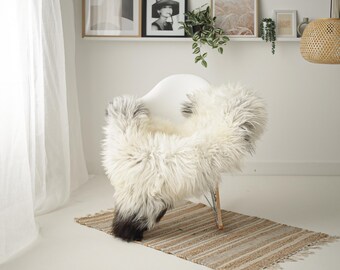 May include: A white sheepskin rug draped over a white chair with a wooden frame. The rug has a black and white pattern and is fluffy. The chair is in a room with white walls and a white curtain.