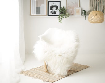 May include: A white faux fur throw draped over a white chair with a wooden base. The chair is sitting on a woven rug in a white room with a window and a bamboo pendant light.