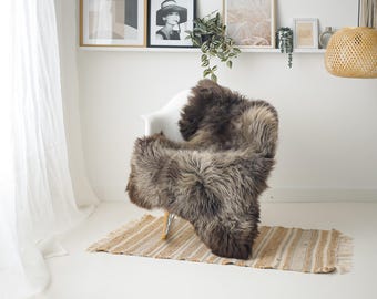Può includere: Un tappeto in pelle di pecora marrone e grigio drappeggiato su una sedia bianca. Il tappeto poggia su un tappeto a righe beige e bianco. La stanza ha un'estetica minimalista con opere d'arte incorniciate e una lampada a sospensione intrecciata.