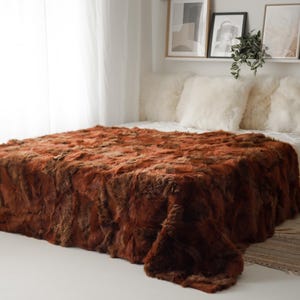 Peut inclure: Un lit avec une grande couverture en fausse fourrure de couleur rouille. Le lit a des oreillers blancs et un drap blanc. Une suspension tissée est suspendue au plafond. La pièce a une palette de couleurs neutres.