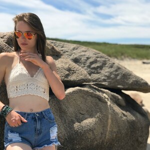 May include: A woman wearing a white crochet halter top, blue denim shorts, and sunglasses. She is standing in front of a large rock.