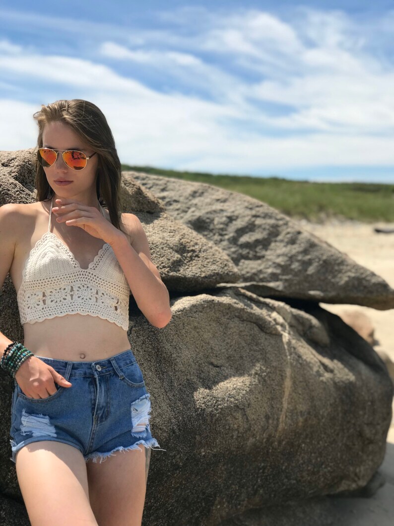 May include: A woman wearing a white crochet halter top, blue denim shorts, and sunglasses. She is standing in front of a large rock.