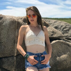 May include: A woman wearing a white crochet top and denim shorts. She is standing in front of a large rock and wearing sunglasses.