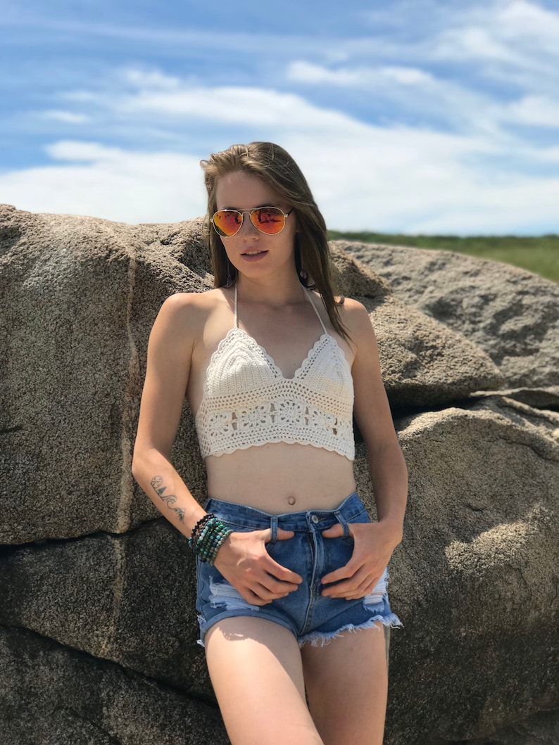 May include: A woman wearing a white crochet top and denim shorts. She is standing in front of a large rock and wearing sunglasses.