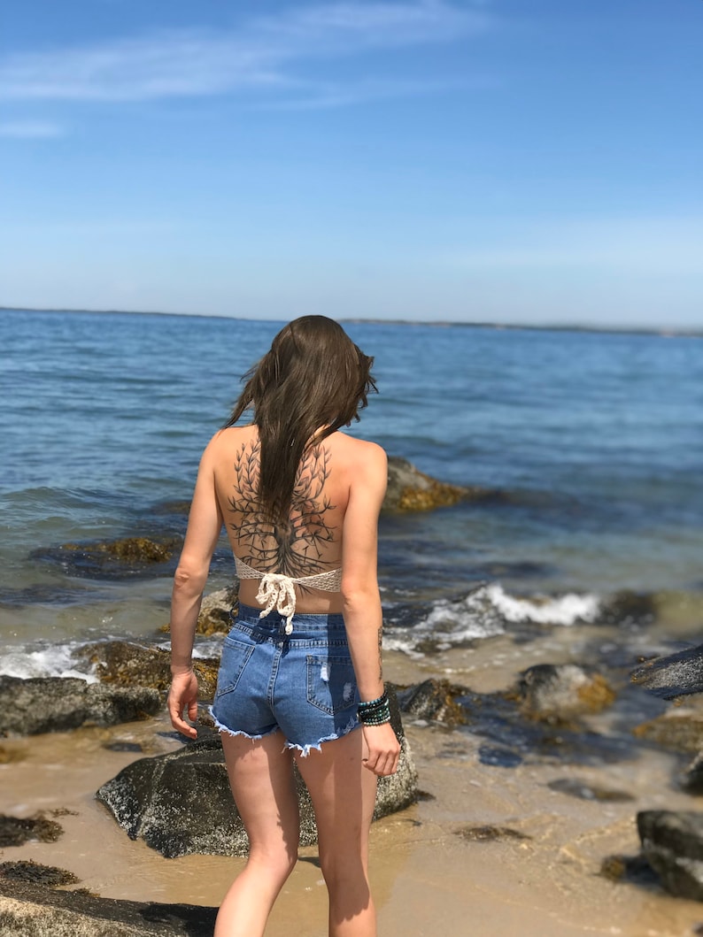 May include: A woman wearing a white crochet top with a tree tattoo on her back, blue denim shorts, and green bracelets, standing on a rocky beach with the ocean in the background.