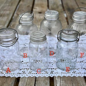 Op de afbeelding: Een verzameling van zes glazen mason jars met metalen deksels en draadsluitingen. De potten variëren in grootte en vorm, sommige hebben reliëfontwerpen. Ze zijn gerangschikt op een wit kanten tafelkleed over een houten oppervlak.