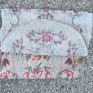 May include: A set of three floral-patterned rugs in muted tones of pink, green, and beige. The rugs are rectangular and semi-circular, with a textured, looped pile. The design features stylized flowers and leaves.