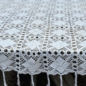May include: A white rectangular tablecloth featuring an intricate crochet pattern with repeating diamond motifs and circular openwork details. The edges of the fabric are finished with a scalloped border and dangling white fringe tassels.