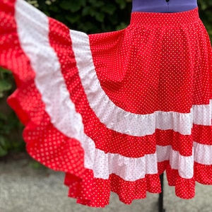May include: A red skirt featuring a white polka dot pattern throughout. The skirt has an elasticized waistband and is designed with alternating horizontal bands of red polka dot fabric and solid white ruffled fabric along the lower section.
