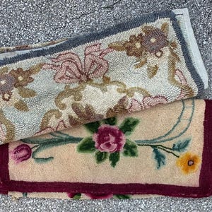 Op de afbeelding: Twee kleine, rechthoekige tapijten met bloemmotieven. Het bovenste tapijt heeft een lichtblauwe achtergrond met roze en bruine bloempatronen. Het onderste tapijt heeft een beige achtergrond met een grote roze roos en groene bladeren.