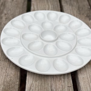 May include: A circular white ceramic serving platter featuring a central round compartment surrounded by two concentric rings of oval-shaped indentations designed for holding individual eggs. The platter has a smooth, glossy finish.
