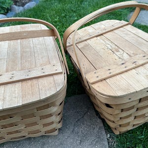 May include: Two light brown picnic baskets with wooden lids and woven sides. Each basket has a curved wooden handle. The lids are made of wooden planks secured with wooden strips.