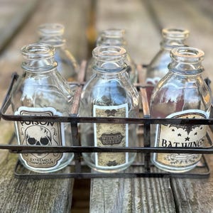 May include: Six clear glass bottles with labels in a metal carrier. The labels read "Poison", "Witches Butter", and "Bat Wings". The bottles are displayed on a wooden surface, evoking a Halloween theme.