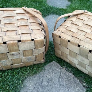 May include: Two woven picnic baskets with tan leather handles. The baskets are made of light brown wood strips woven together in a rectangular pattern. The baskets are sitting on green grass and a stone.