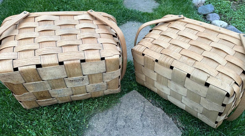 May include: Two woven picnic baskets with tan leather handles. The baskets are made of light brown wood strips woven together in a rectangular pattern. The baskets are sitting on green grass and a stone.