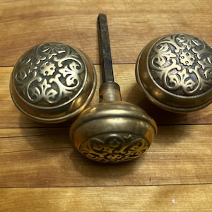 May include: Three antique brass door knobs with ornate floral designs. The knobs are a warm gold color and sit on a wooden surface. The center knob has a long, thin metal rod attached.