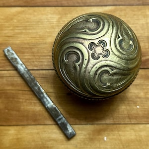May include: An antique, spherical metal container with ornate, swirling patterns and a small, rectangular metal tool. The container is gold-toned with a detailed design. The tool is silver-colored and lies next to the container on a wooden surface.