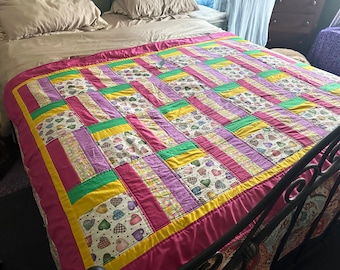 May include: A vibrant patchwork quilt in shades of pink, purple, yellow, and green, with some blocks featuring heart patterns. The quilt is laid on a bed with a neutral-toned duvet cover. The quilt's dimensions are not visible.
