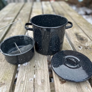 May include: A black enamel cooking pot set with white speckles. The set includes a pot with two handles, a lid, and a small divided bowl with a metal handle. The items are on a wooden surface.