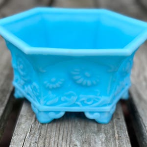 May include: A bright turquoise blue hexagonal bowl made of pressed glass. The exterior features a raised relief pattern of stylised flowers and scrolling vines. The bowl is supported by small, decorative feet at each of the six bottom corners.