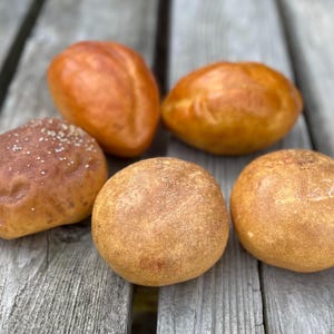 May include: An overhead shot of five freshly baked rolls. The rolls vary in shape and color, ranging from golden brown to a darker, reddish-brown hue. Some are sprinkled with seeds, while others have a smooth, glossy surface. The rolls are arranged on a weathered wooden surface.