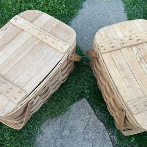May include: Two light brown, rectangular picnic baskets with wooden lids. The lids are secured with wooden slats and metal rivets. Each basket has a woven body and a leather handle.