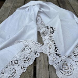 Op de afbeelding: Witte linnen tafelkleed met ingewikkelde kanten rand. Het tafelkleed is gedrapeerd en toont de delicate kanten rand. De stof lijkt zacht en licht gekreukt, wat wijst op een vintage of handgemaakte kwaliteit. Het kantontwerp heeft bloempatronen.