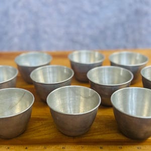 May include: A collection of ten small, silver-colored metal bowls arranged on a wooden surface. The bowls have a rounded shape with a slightly flared rim. The background is a blurred, light blue color.
