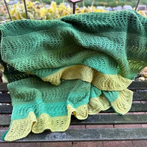 May include: A handmade, knitted blanket in shades of green and yellow. The blanket features a textured pattern and is draped over a wooden bench. The color palette includes dark green, light green, and yellow, creating a cozy and inviting feel. Perfect for home decor.