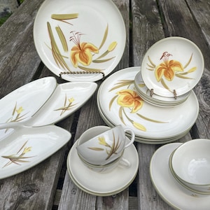 May include: A collection of off-white ceramic dinnerware featuring a painted yellow lily and green leaf motif. The set includes large dinner plates, smaller side plates, a divided serving dish, and several teacups with matching saucers.