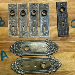 May include: A collection of antique door hardware, featuring five rectangular plates with floral and geometric patterns, two oval plates with ornate edges, and one dark rectangular plate. The hardware is metal and has a vintage appearance.