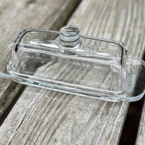 May include: A clear glass butter dish, rectangular in shape, with a matching lid and a small, round handle. The dish is displayed on a weathered wooden surface.