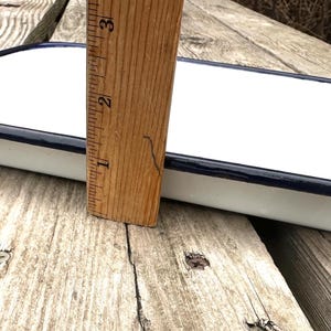 May include: A rectangular white enamelware tray with a dark blue rim. A wooden ruler is placed next to the tray, showing a depth of approximately 1 inch. The tray is on a weathered wooden surface.