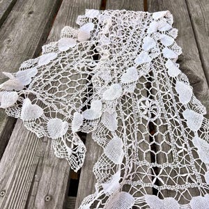 Puede incluir: Una delicada bufanda de encaje blanco roto con un patrón floral y geométrico. La bufanda está colocada sobre una superficie de madera envejecida, mostrando su intrincado diseño calado y bordes festoneados. El encaje presenta detalles en forma de corazón.