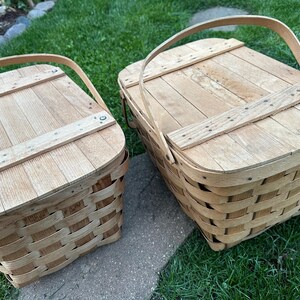 May include: Two light brown picnic baskets with wooden lids and handles. The baskets are made of woven strips of wood and have a rectangular shape. The lids are made of wooden planks and are attached with small nails. The baskets are sitting on a stone surface and green grass.
