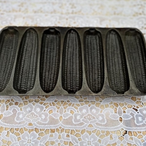 May include: A cast iron cornbread pan with six corn cob molds. The pan is dark gray and has handles on each end. The pan sits on a white lace tablecloth.