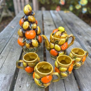 Puede incluir: Un juego de té de cerámica con una tetera, cuatro tazas y un centro de mesa decorativo con forma de fruta. El juego está adornado con un patrón de naranjas, limones y hojas verdes. Las piezas están dispuestas sobre una mesa de madera envejecida, mostrando una estética vintage.
