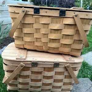 May include: Two stacked, light brown woven picnic baskets with wooden handles and hinged lids. The baskets have a classic rectangular shape and are made of woven wood strips. The handles are attached with metal hinges.