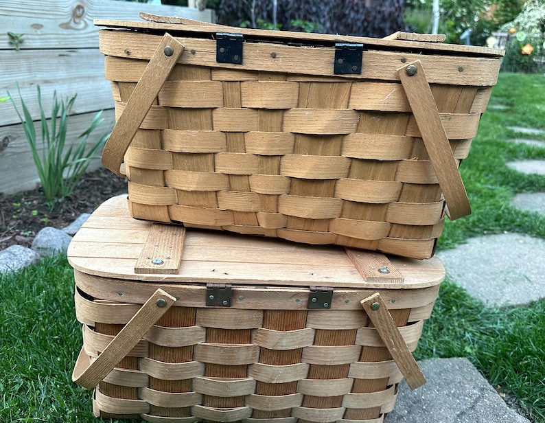 May include: Two stacked, light brown woven picnic baskets with wooden handles and hinged lids. The baskets have a classic rectangular shape and are made of woven wood strips. The handles are attached with metal hinges.